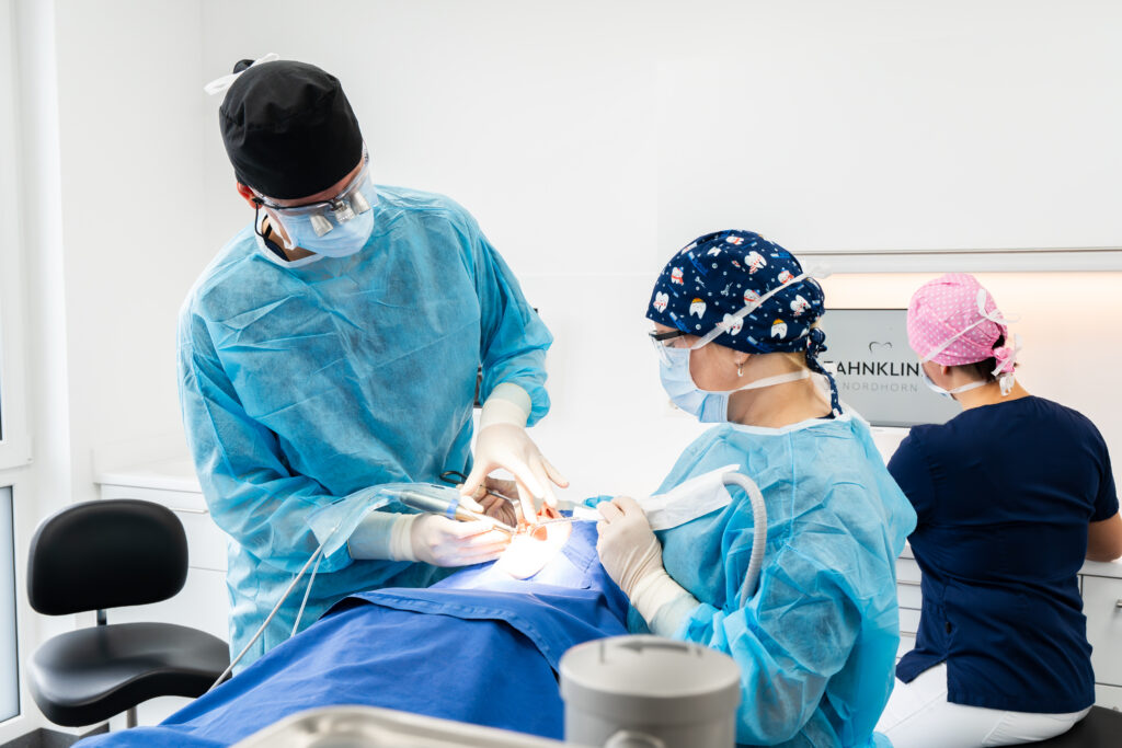 Zahnärztliches Team in blauen OP-Kitteln bei einer Behandlung im Behandlungsraum, mit OP-Maske und hellen Flächen, Zahnklinik Nordhorn im Hintergrund.