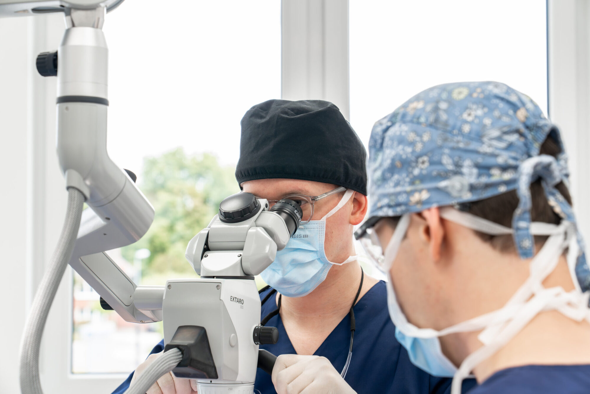 Präzise Zahnarztbehandlung unter dentalem Mikroskop mit Schutzbrille, OP-Maske und blauer Kopfbedeckung; am Gerät EXTARO.