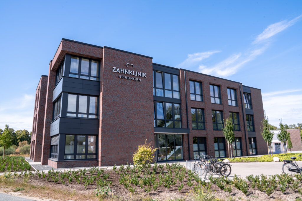 Außenansicht der Zahnklinik Nordhorn – modernes Backsteingebäude mit großen Glasfronten unter blauem Himmel. Das Bild zeigt den Eingangsbereich und die gepflegte Außenanlage der Praxis.