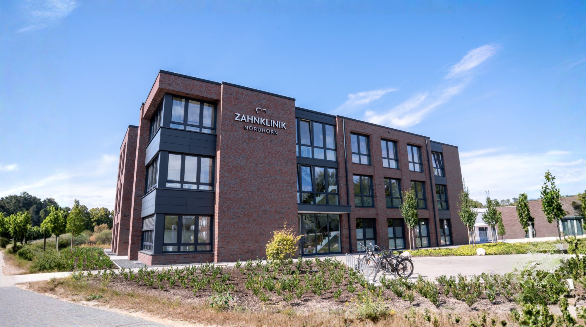 Moderne Außenansicht der Zahnklinik Nordhorn mit roter Klinkerfassade, großen Glasfronten und gepflegtem Eingangsbereich – Zahnmedizin auf höchstem Niveau in Nordhorn.