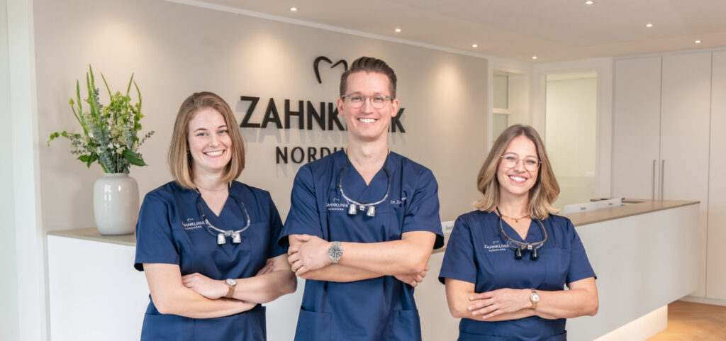 Drei Zahnärztinnen und Zahnärzte der Zahnklinik Nordhorn stehen gemeinsam im Empfangsbereich der Praxis. Sie tragen blaue Arbeitskleidung und Lupenbrillen um den Hals und lächeln in die Kamera. Im Hintergrund ist das Praxislogo „Zahnklinik Nordhorn“ zu sehen.