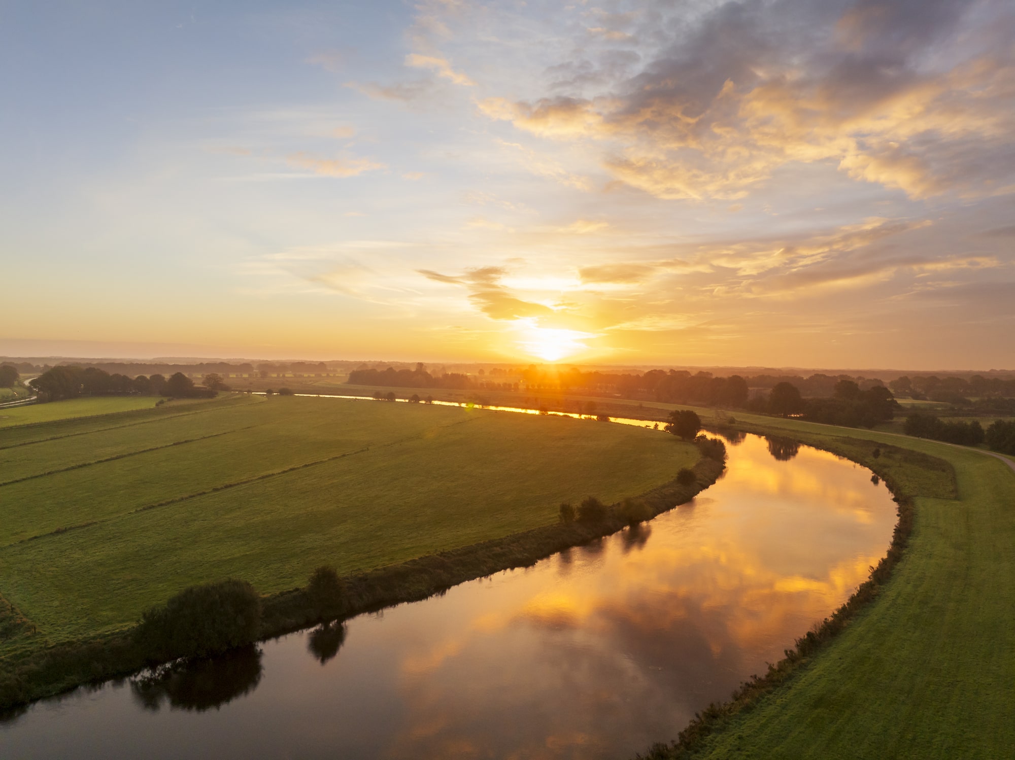 Luftaufnahme einer Flusslandschaft bei Sonnenuntergang. Der Fluss windet sich durch grüne Wiesen und Felder, während sich das goldene Licht des Himmels im Wasser spiegelt.