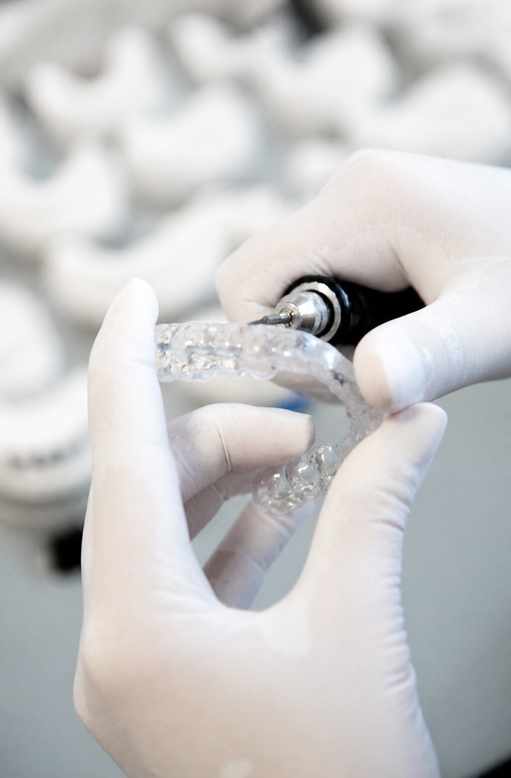 Gummihandschuh-Hände halten eine transparente Zahnschiene und ein feines dentales Bearbeitungswerkzeug.